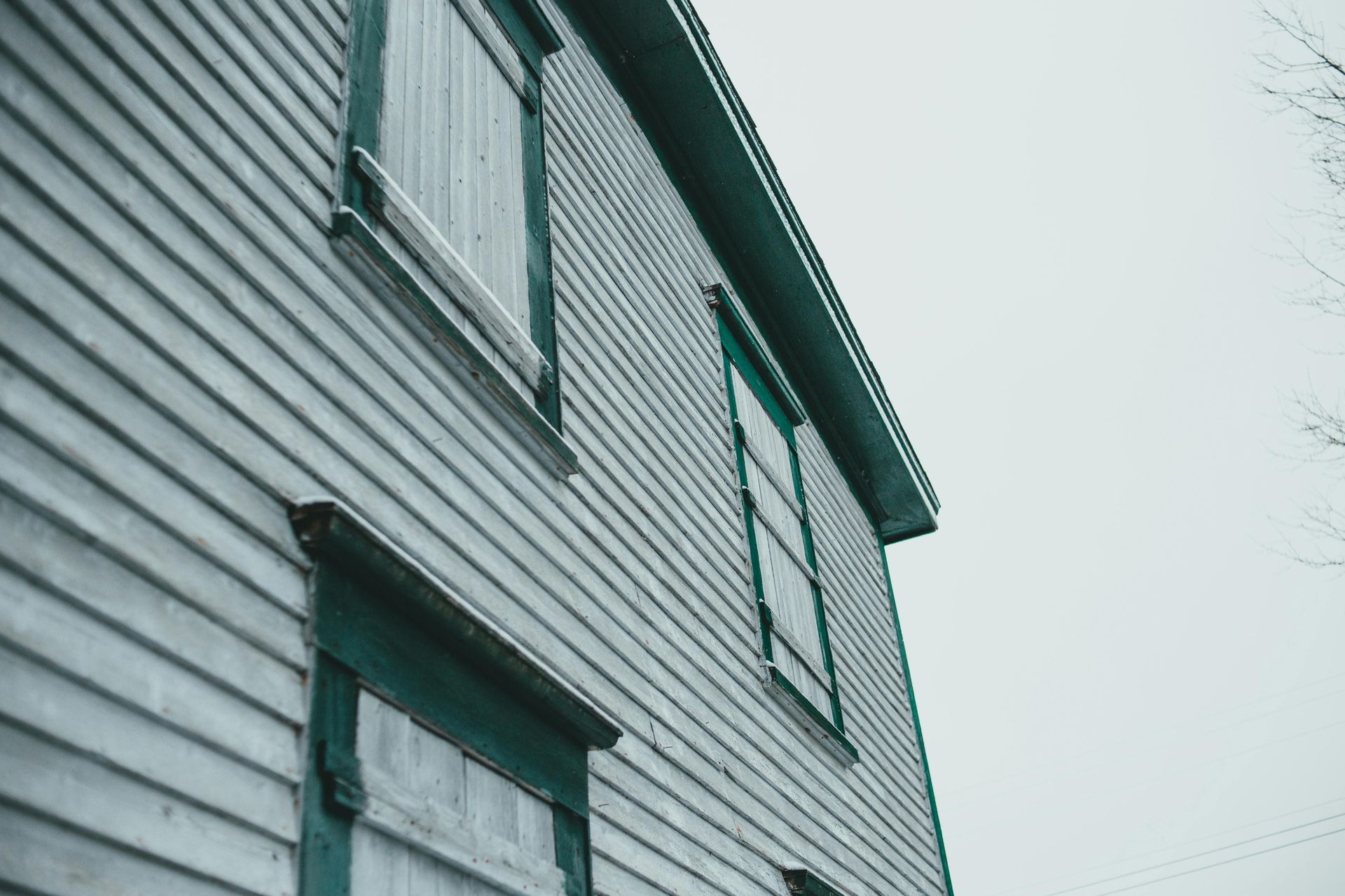 Side view of an old, weathered house with boarded-up windows. Gray siding, teal trim, overcast sky.