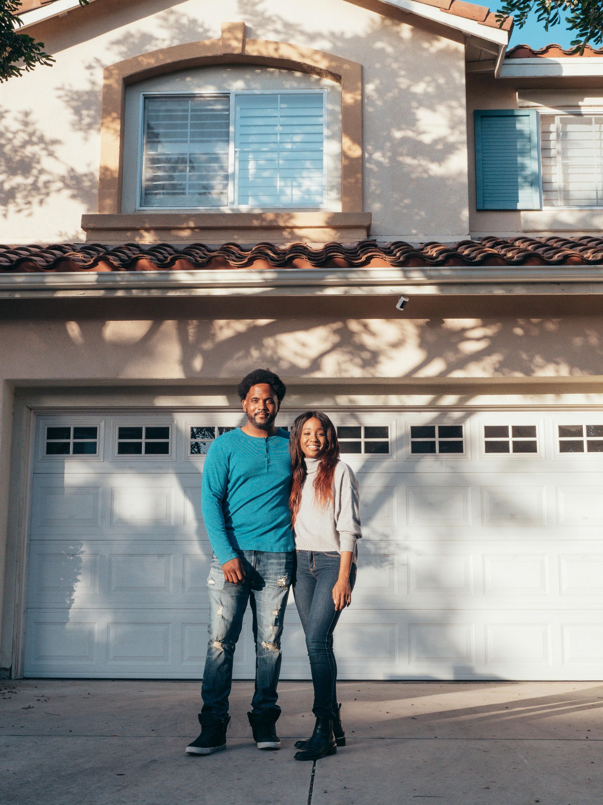 Two people standing in front of a garage door at a suburban house, one in a teal shirt and one in a white top.