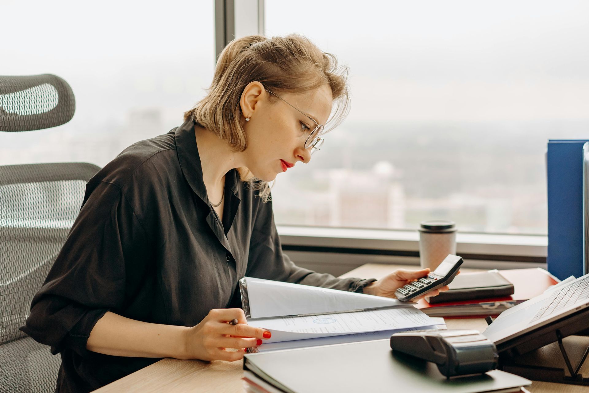 Woman in glasses calculates with papers and a calculator at a desk near a window.