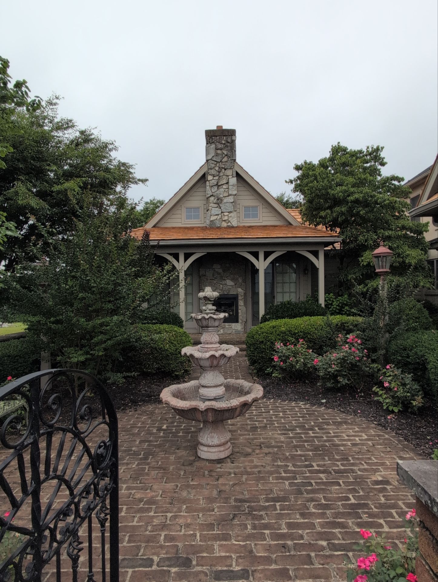 Brick courtyard with a small stone fountain, garden shrubs, and a cottage-style house in the background
