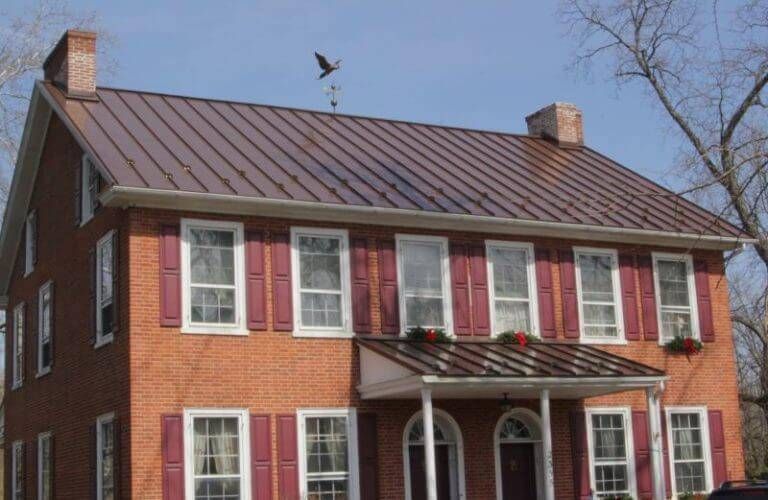 A two-story brick house with a brown metal roof, red shutters, and a front porch, set against a clear blue sky.
