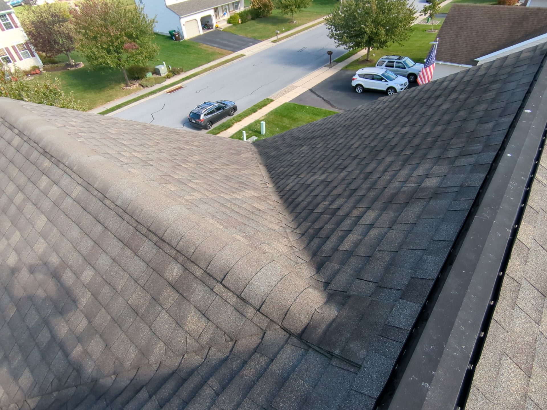 View from a rooftop over a suburban street, with gray shingles, parked cars, and a house with an American flag.