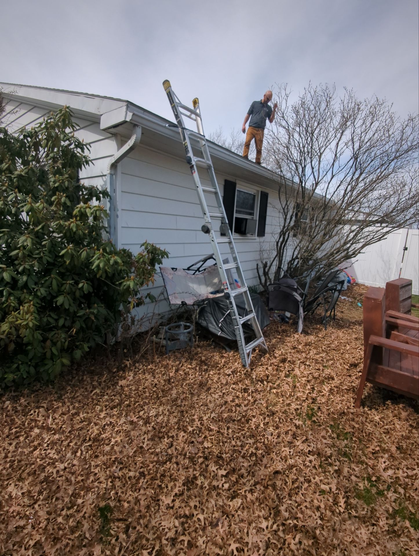 Worker on ladder repairing a white house roof in a leaf-covered yard, with another worker on the roof.