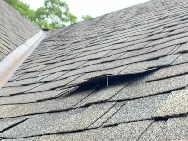Damaged asphalt shingles on a roof with a hole. Gray shingles with visible wear and tear.
