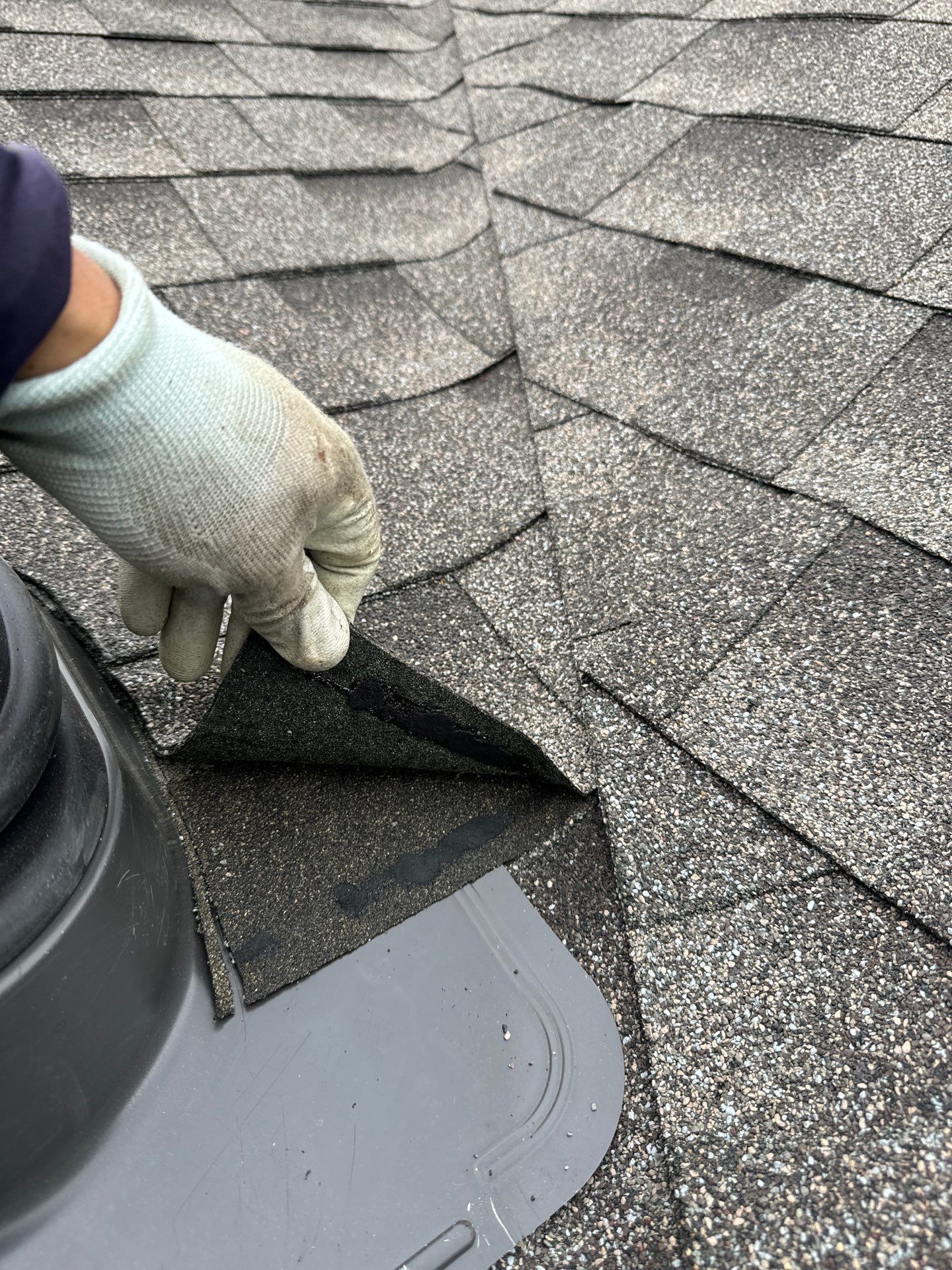Gloved hand lifting a damaged shingle on a gray asphalt roof near a roof vent