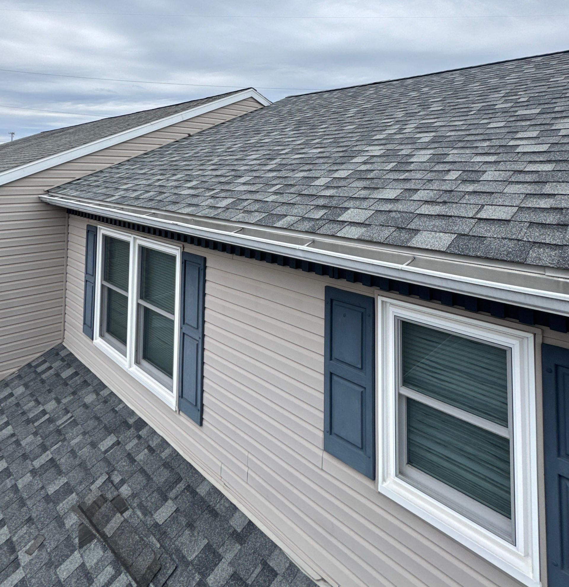 Aerial view of a gray shingled roof and beige house exterior with several windows.