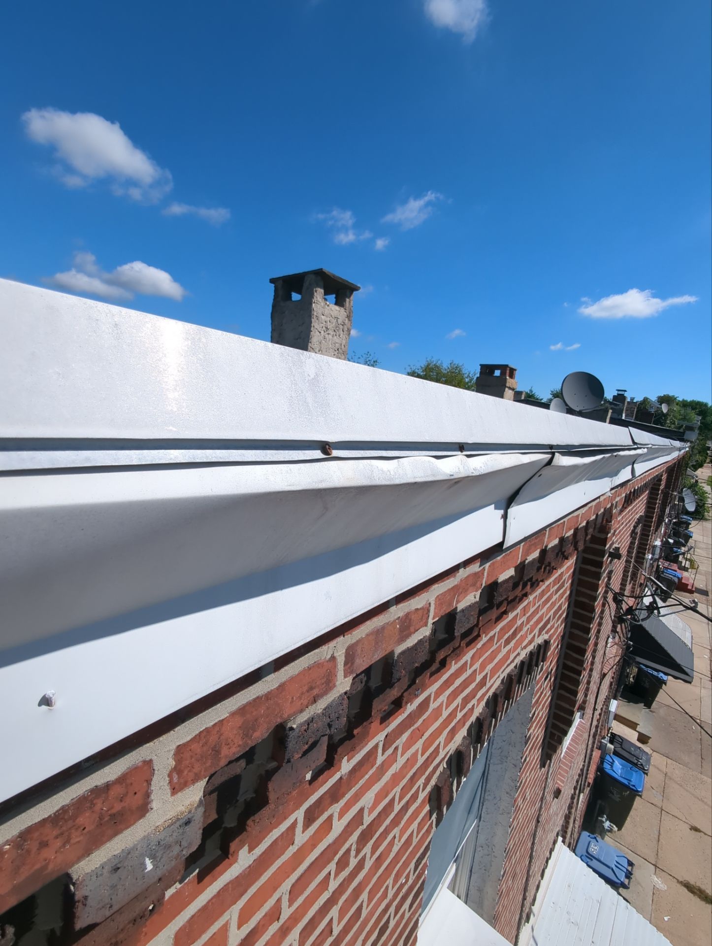 Rooftop with flat black roofing, white vents, and a damaged red-brick wall beside trees and nearby houses