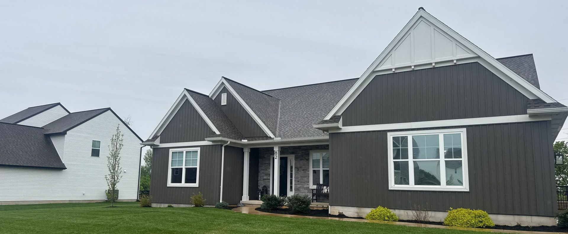 Modern gray house with white trim and dark roof on a green lawn under cloudy skies