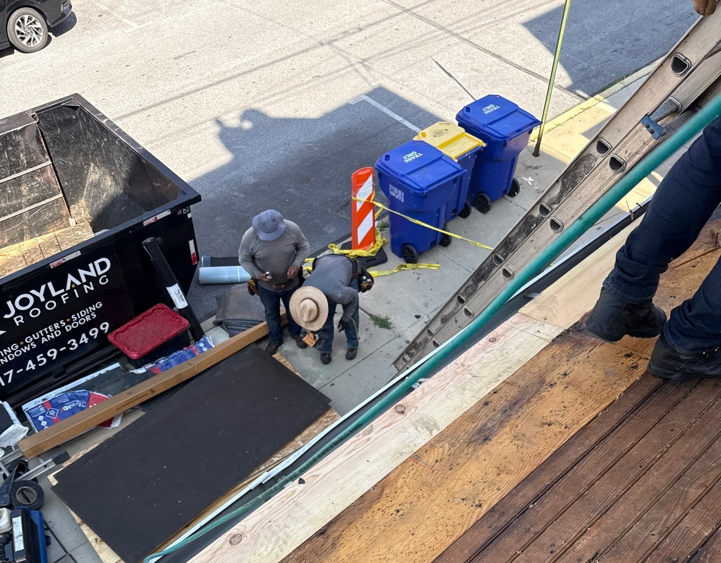 Workers near blue bins and a dumpster beside a sidewalk, viewed from above.