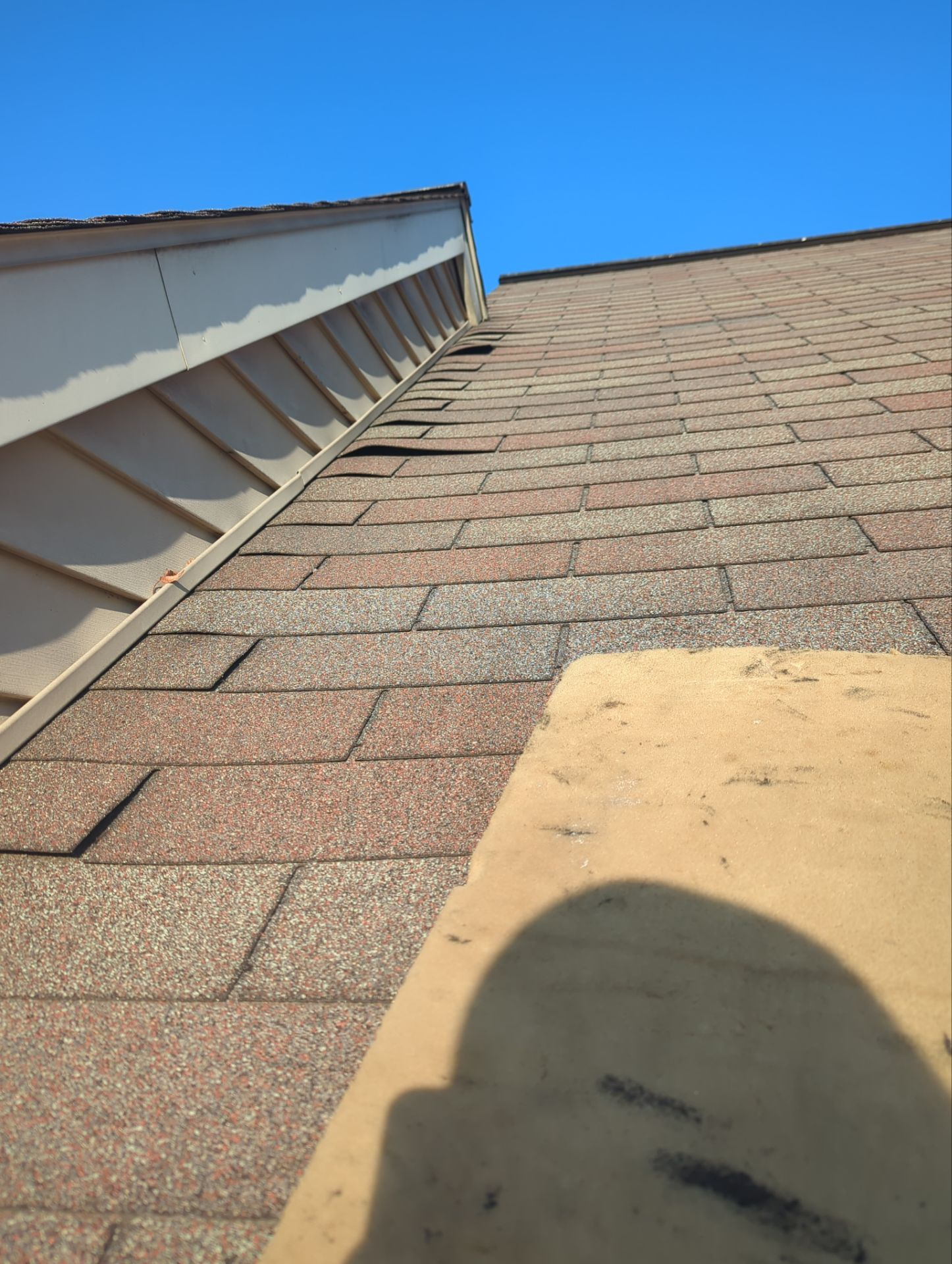 Steep shingled roof beside white siding under a clear blue sky, with a shadow in the foreground.