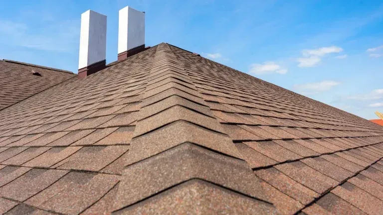 Brown shingle roof with two white chimneys against a blue sky.