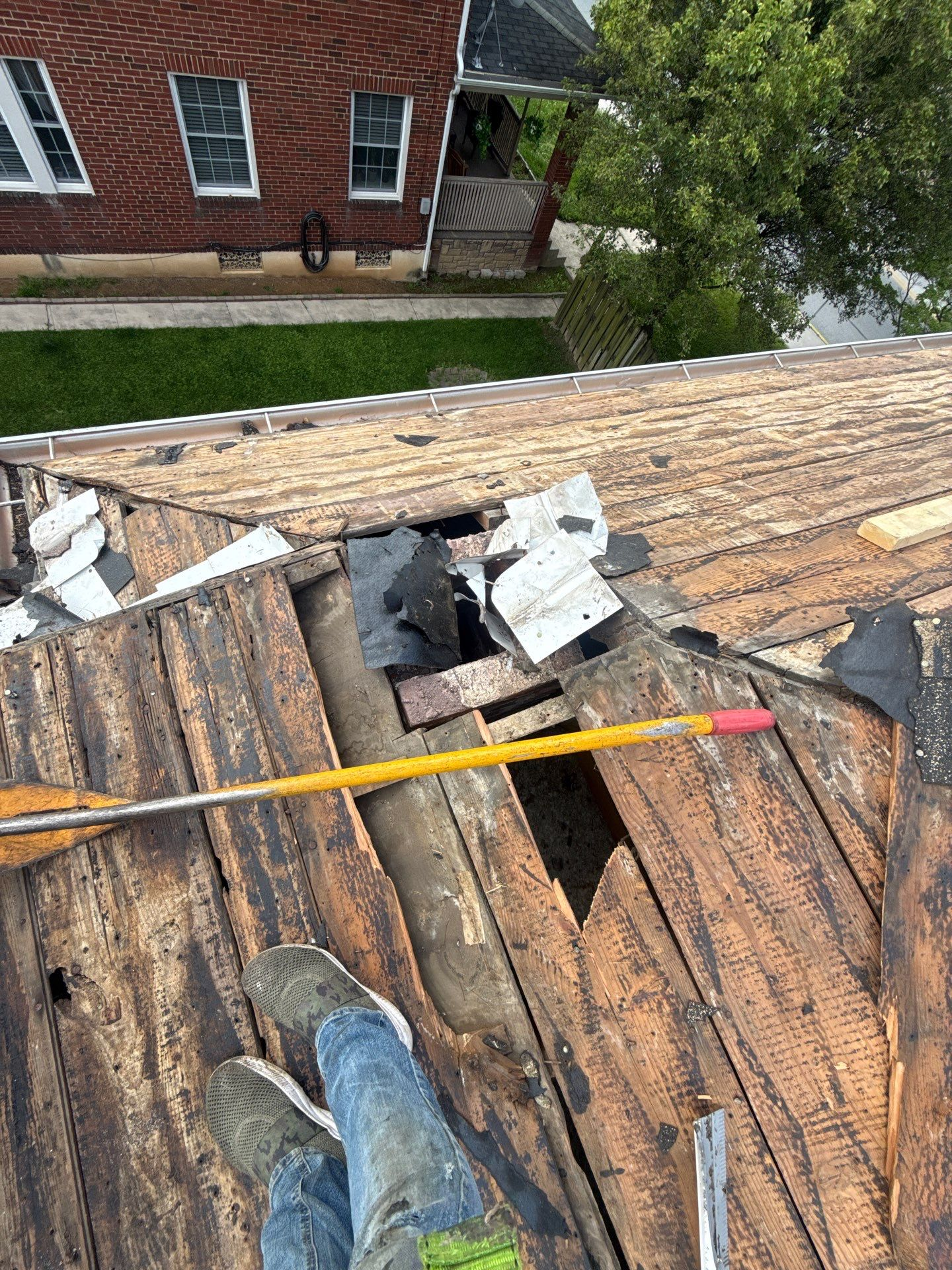 Roof with damaged shingles and scattered debris, viewed from above, with a red brick house in the background