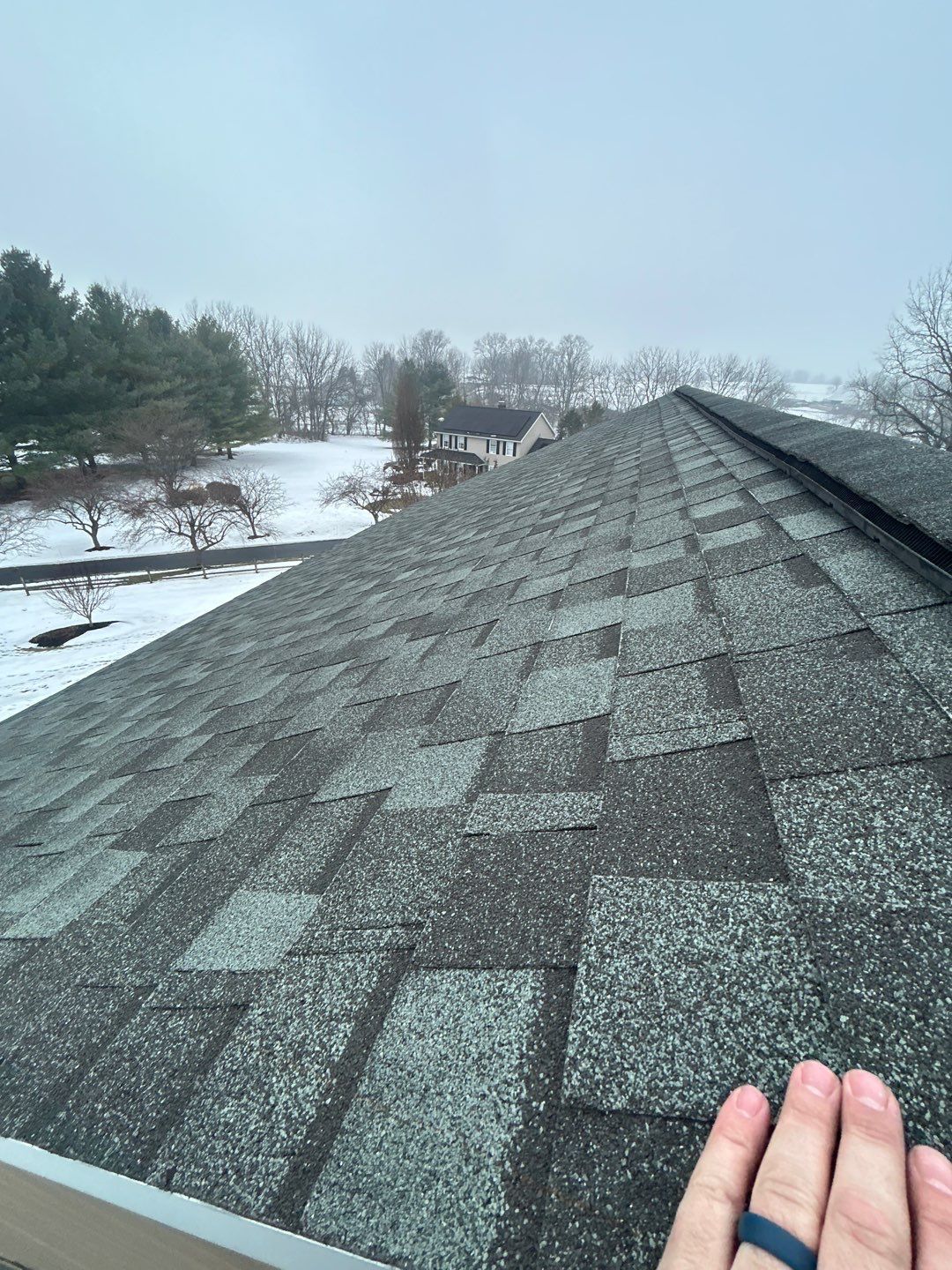 Snowy rooftop with dark shingles, seen from above, with a hand in the lower right corner