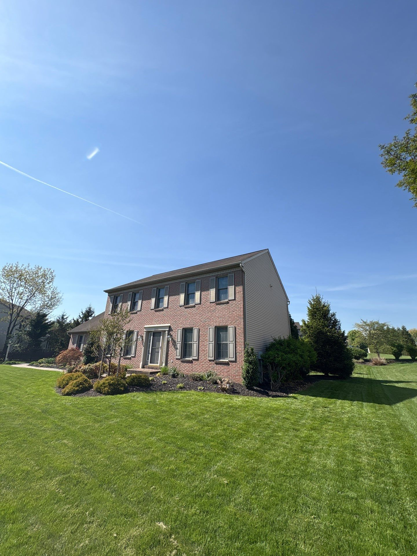 Two-story brick house with green shutters on a sunny lawn under a clear blue sky