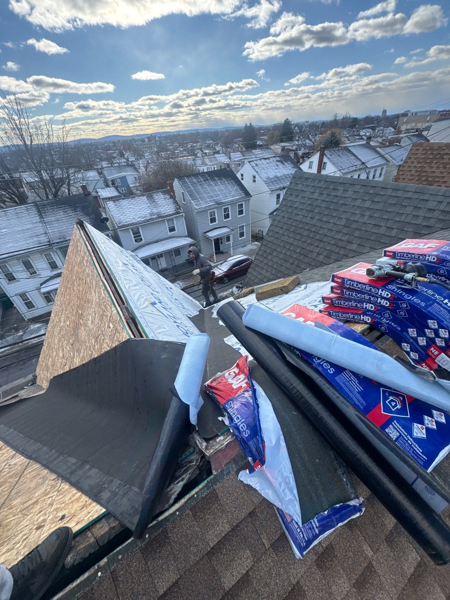 Snow-covered rooftops with roofing materials on a steep roof under a cloudy winter sky