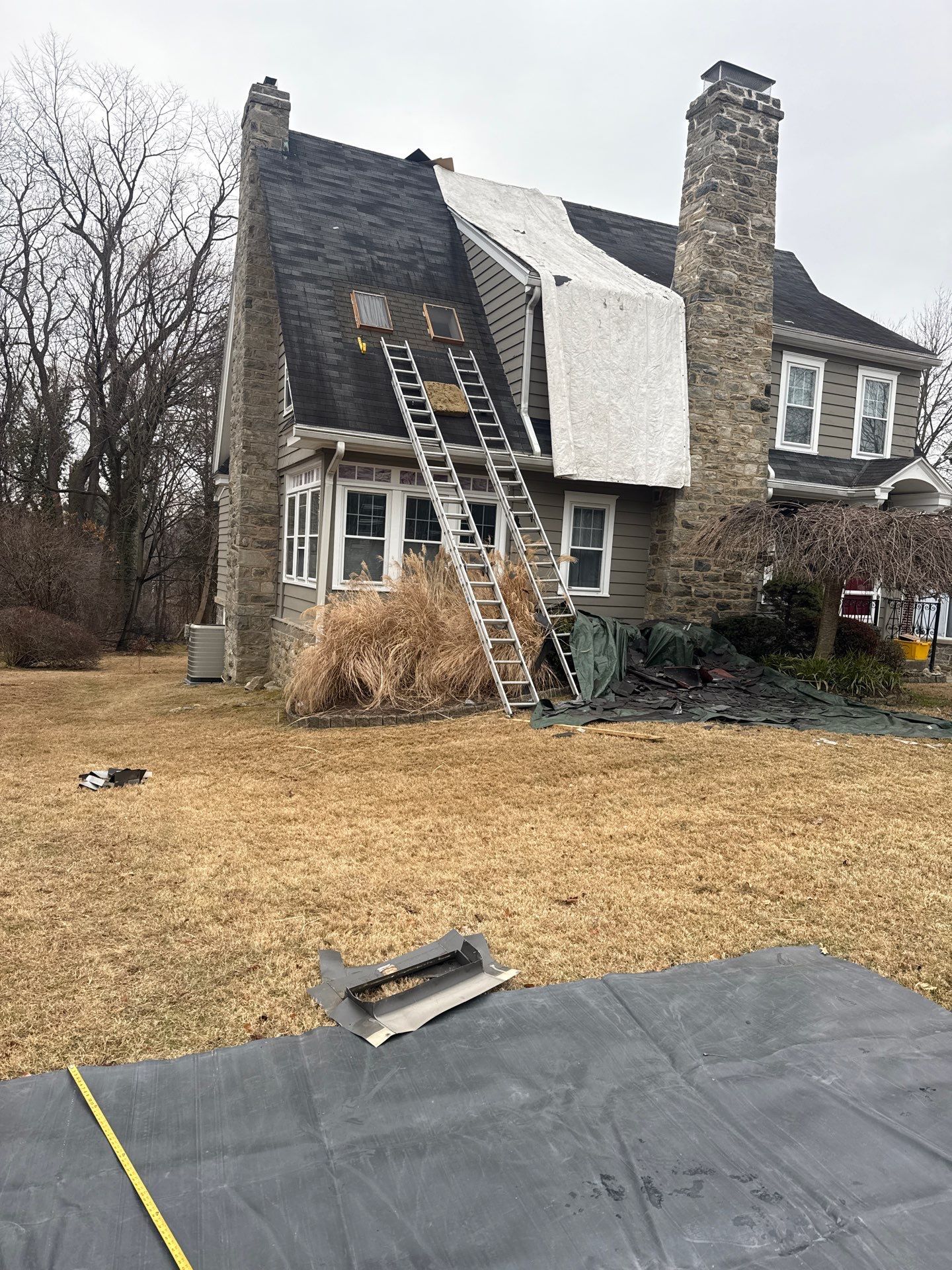 Roof replacement project with exposed roofing materials, ladders, protective covering, and stone chimneys on South Rolling Road in Springfield PA