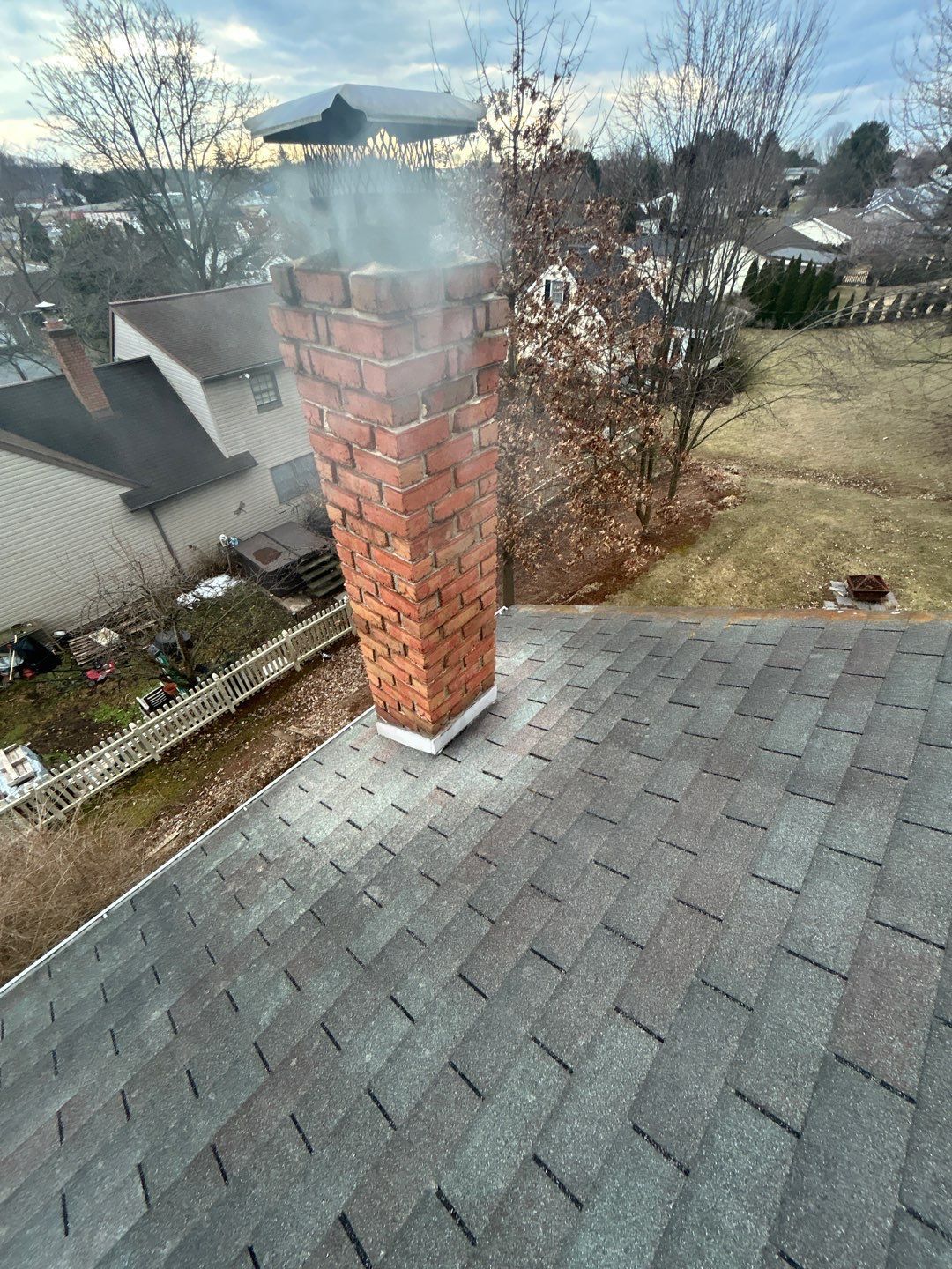 Red brick chimney on a shingled roof with smoke rising, overlooking a suburban neighborhood