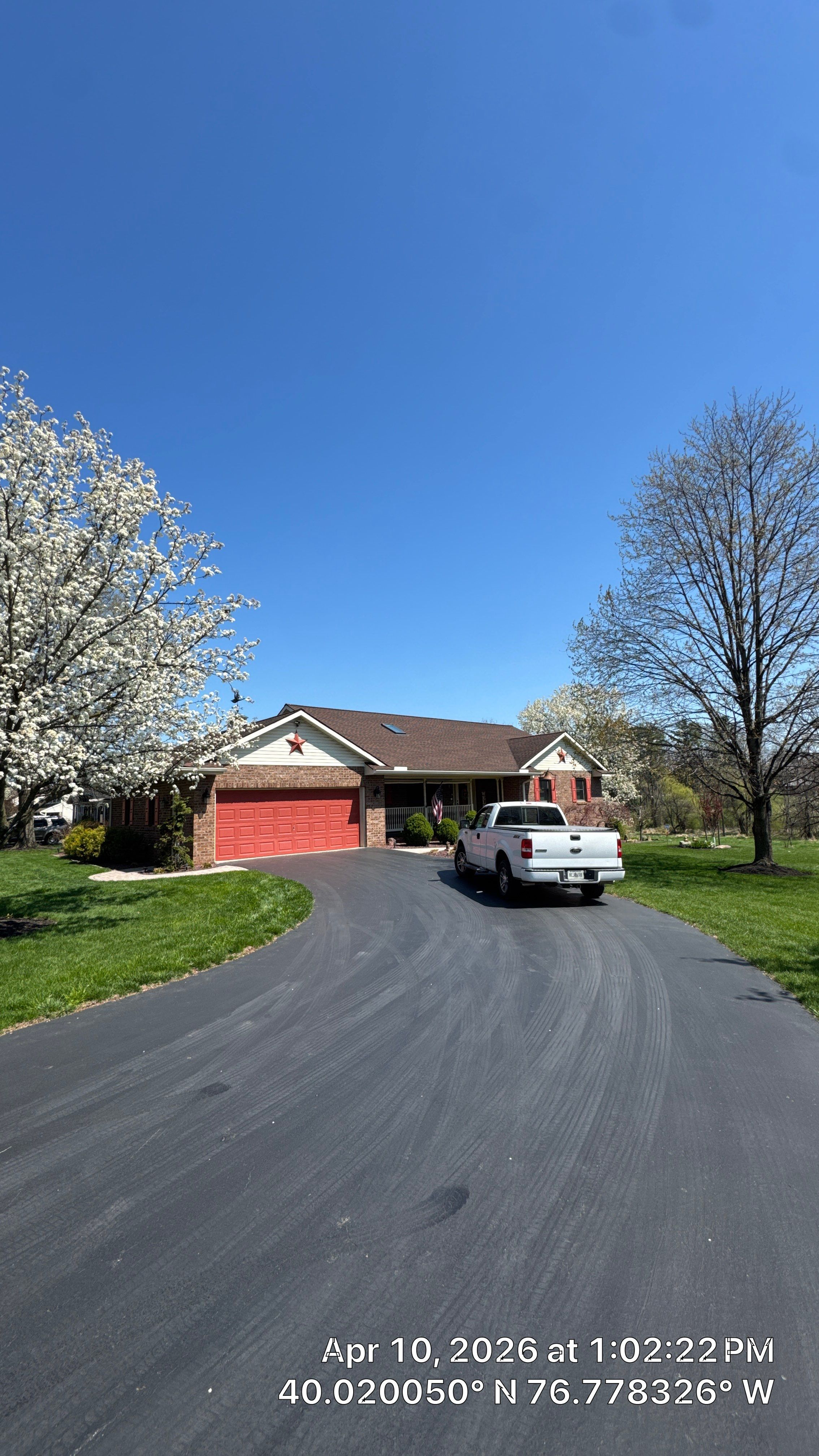 Driveway leading to a red brick house with a white pickup truck and blooming trees under a clear blue sky