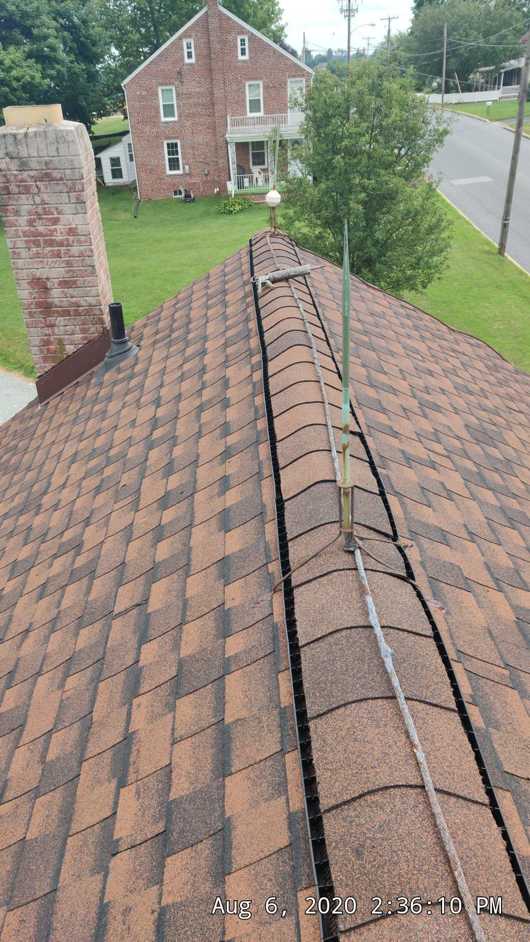 Roof ridge with shingles and a metal vent pipe, viewed from above near a brick house and street.