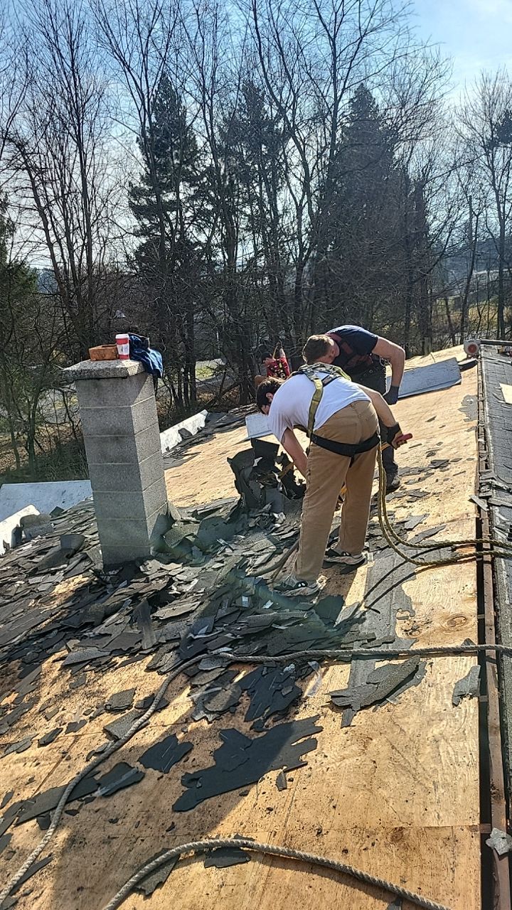 Workers on a rooftop repairing shingles beside a brick chimney on a sunny day.