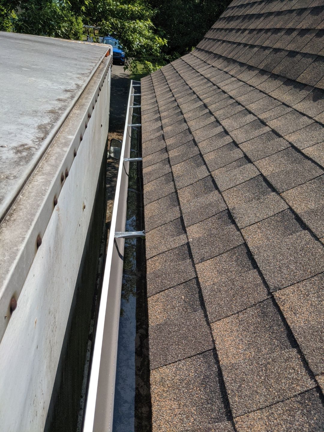 Rooftop gutter between two sloped shingle roofs, with leaves and trees visible below