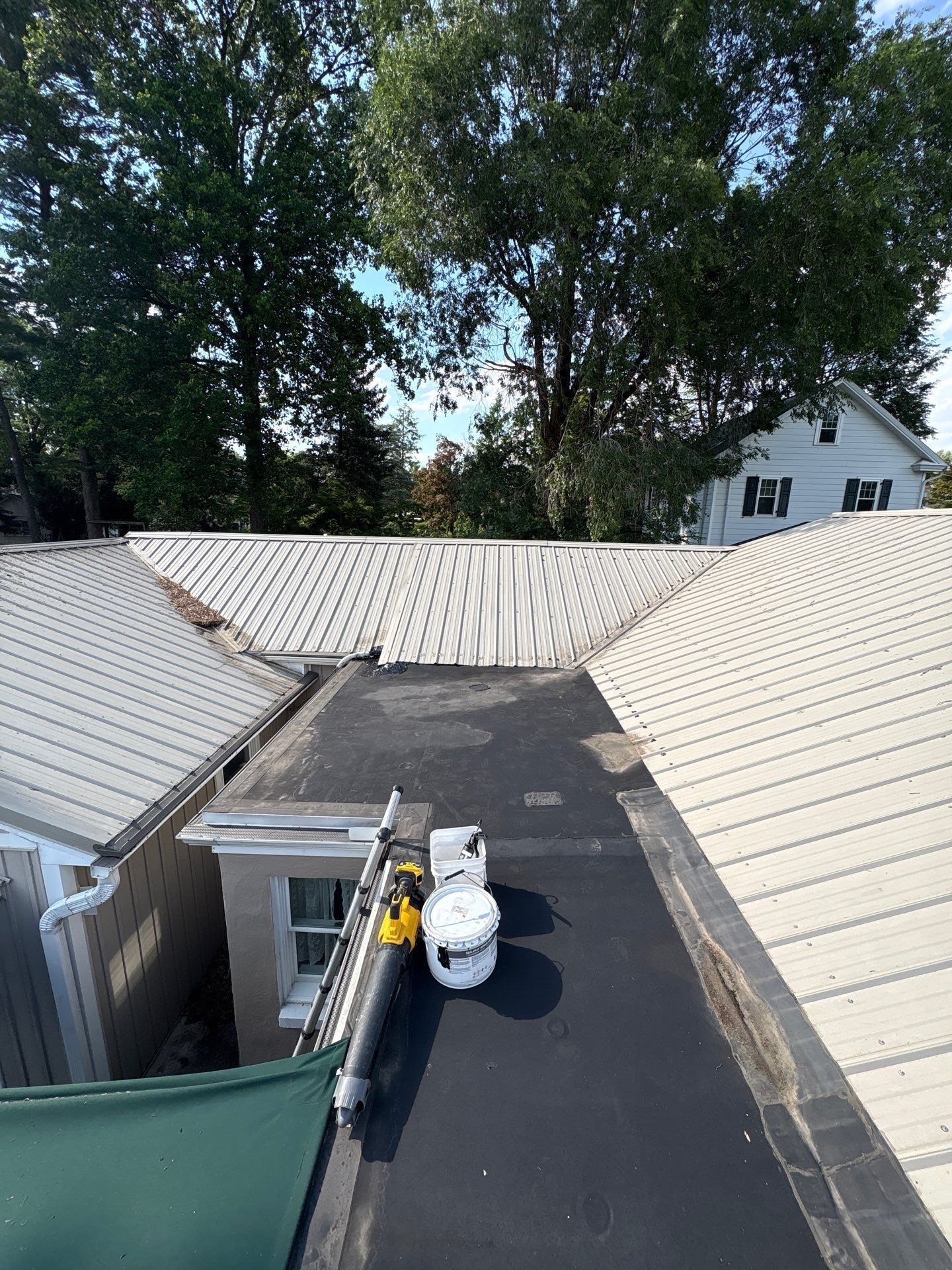 Roof under repair with a black membrane strip between white metal roofs and trees in the background