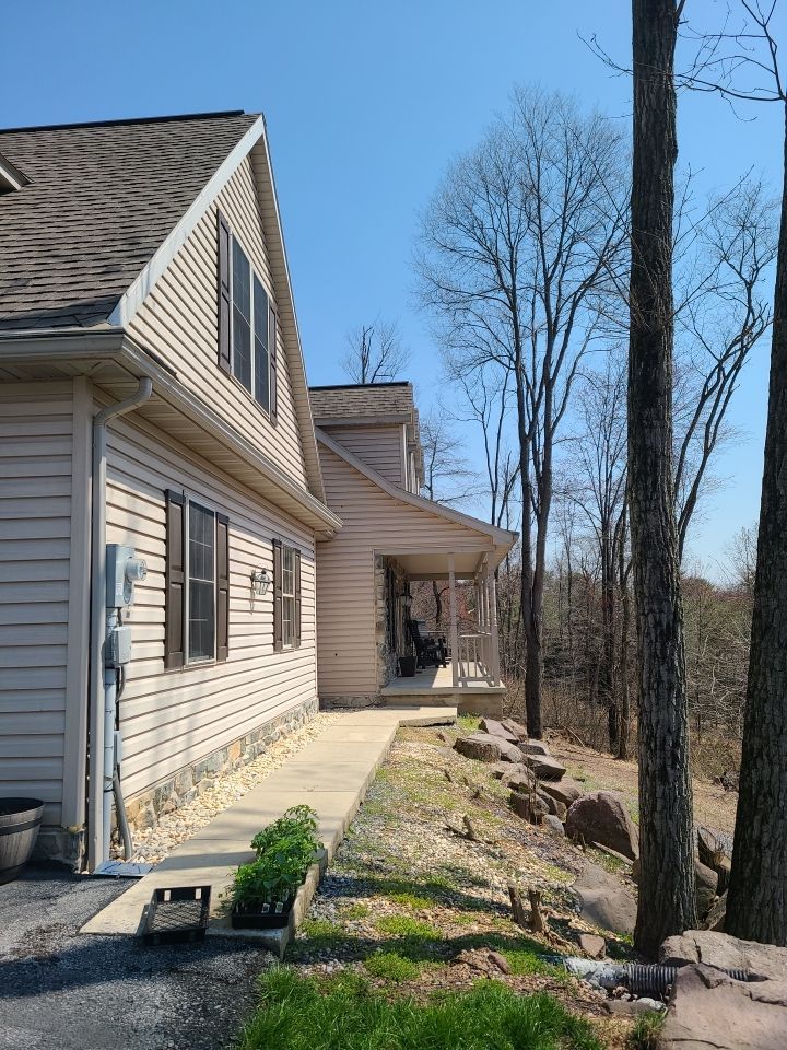 Side of a beige house with a porch, gravel path, and leafless trees under a clear blue sky