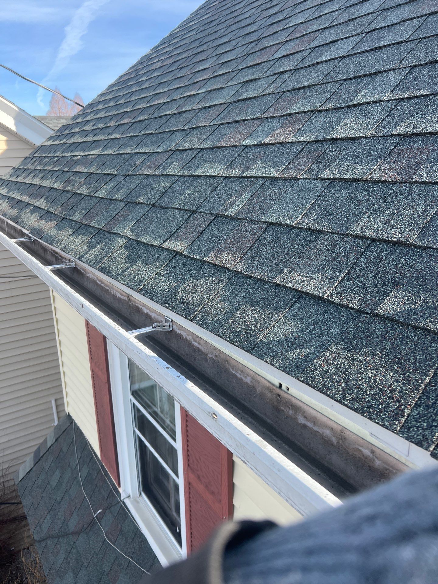Asphalt shingle roof with an open aluminum gutter showing debris buildup and exposed gutter hangers during a roof and drainage inspection.