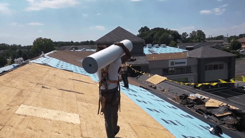 Roofer carrying a roll of material across a partially completed roof on a sunny day.