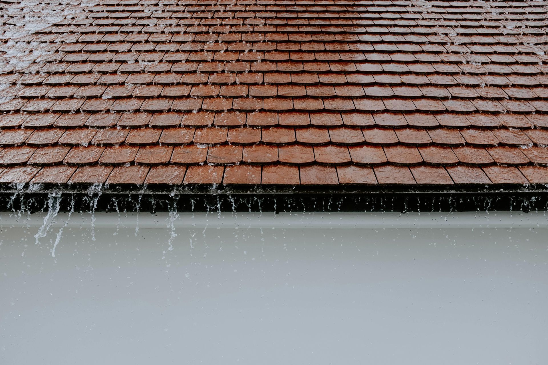 Rain falling off a brown tiled roof into a white gutter.