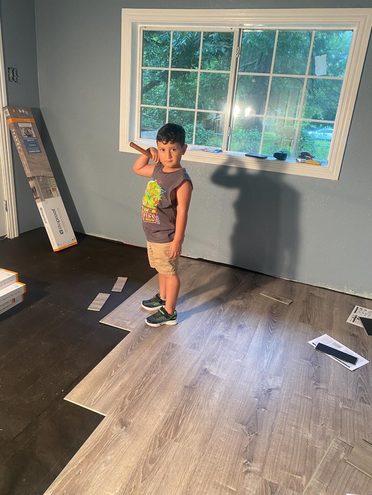 Child standing in a room with partially installed gray flooring, holding a stick, near a window.