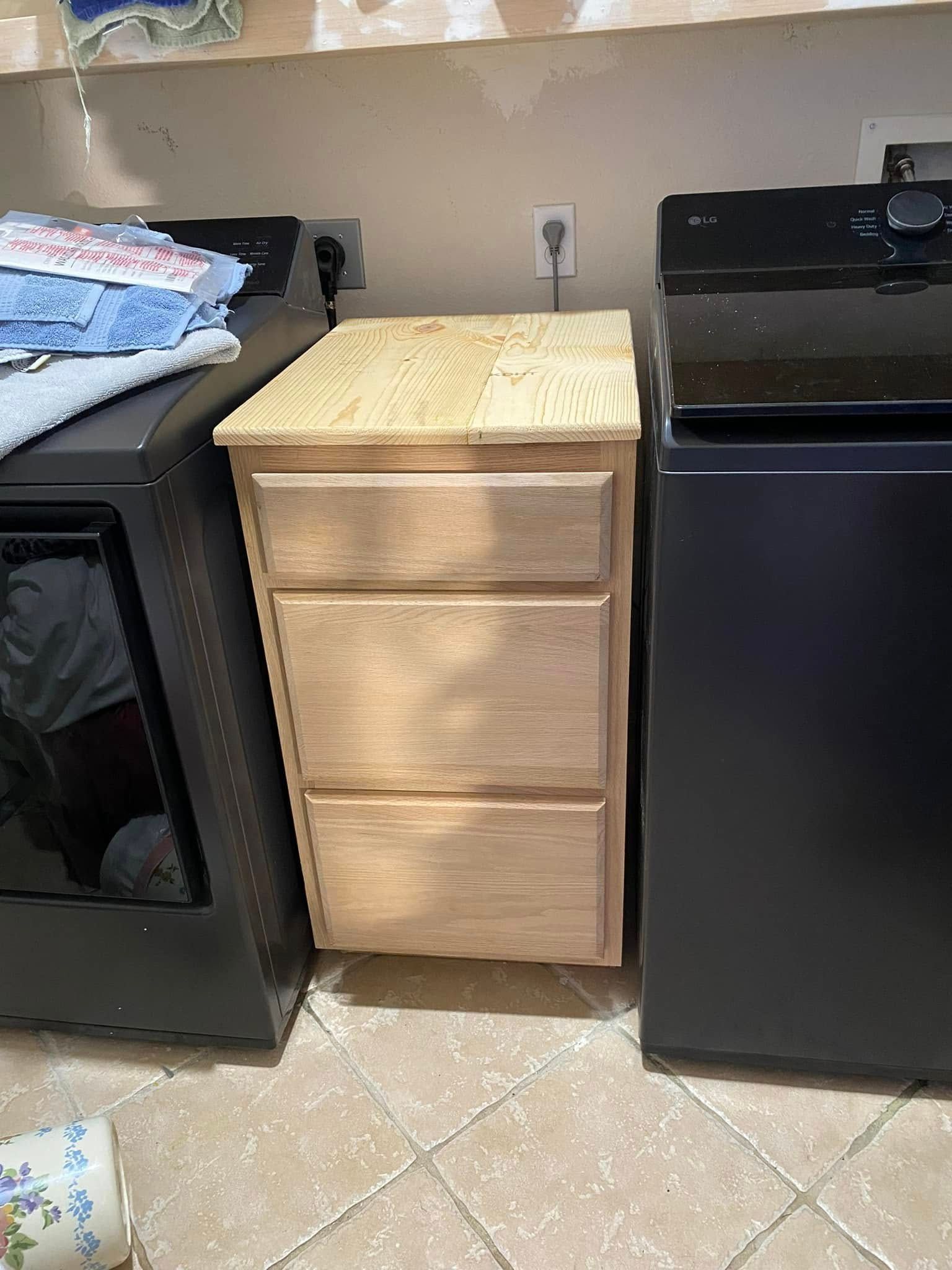 A wooden three-drawer cabinet sits between two black laundry machines in a laundry room.