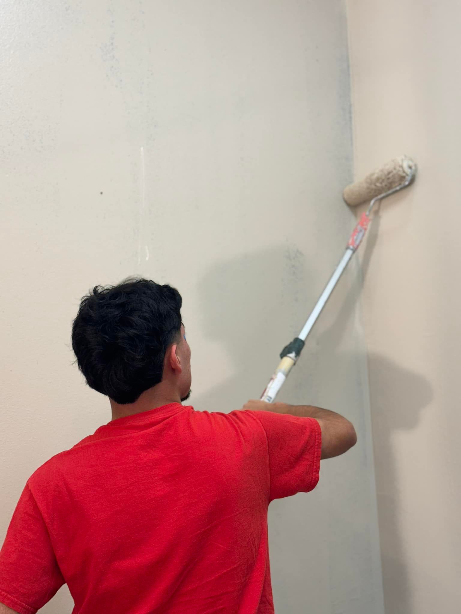 Person in red shirt painting wall with a roller and extendable handle.