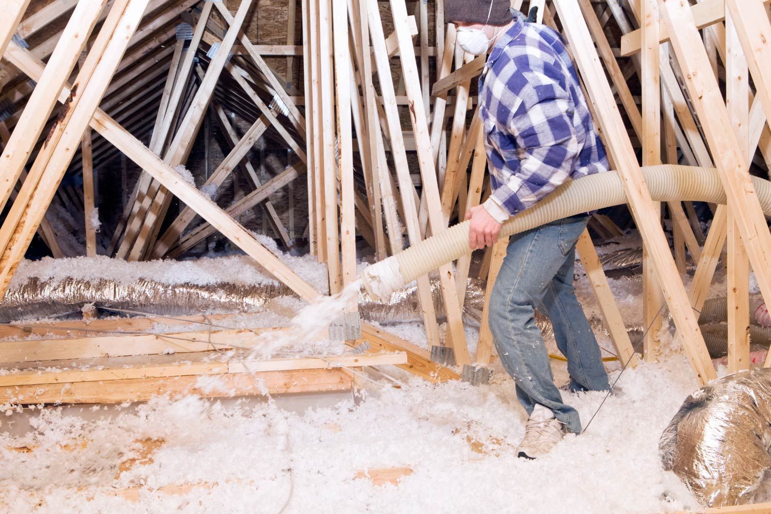 Person in attic blowing insulation with a hose. Wood framing visible; floor covered in loose insulation.