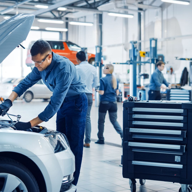 Mechanic working on a white car in a bright auto repair shop, with tools and other workers in the background