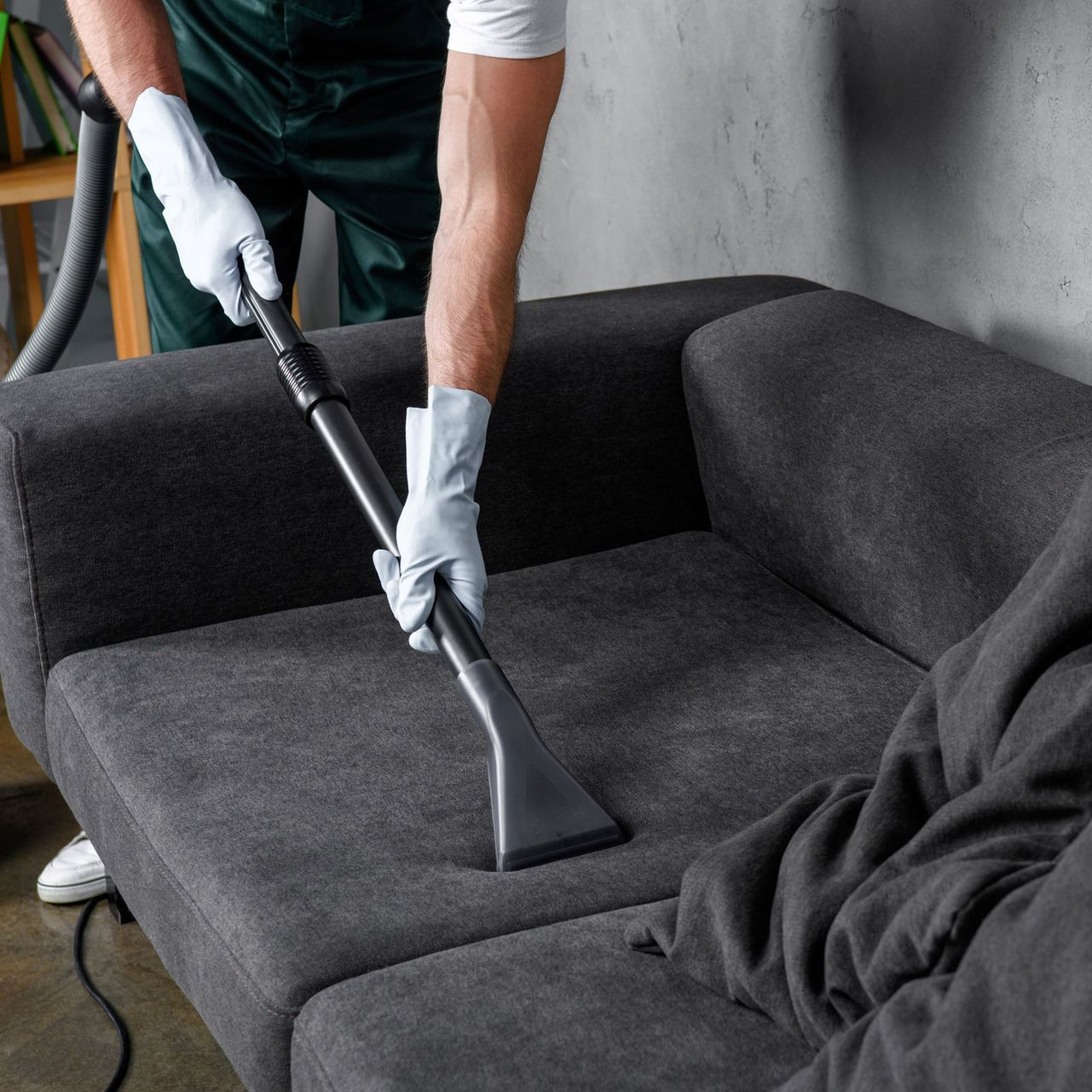 A man is cleaning a black couch with a vacuum cleaner.