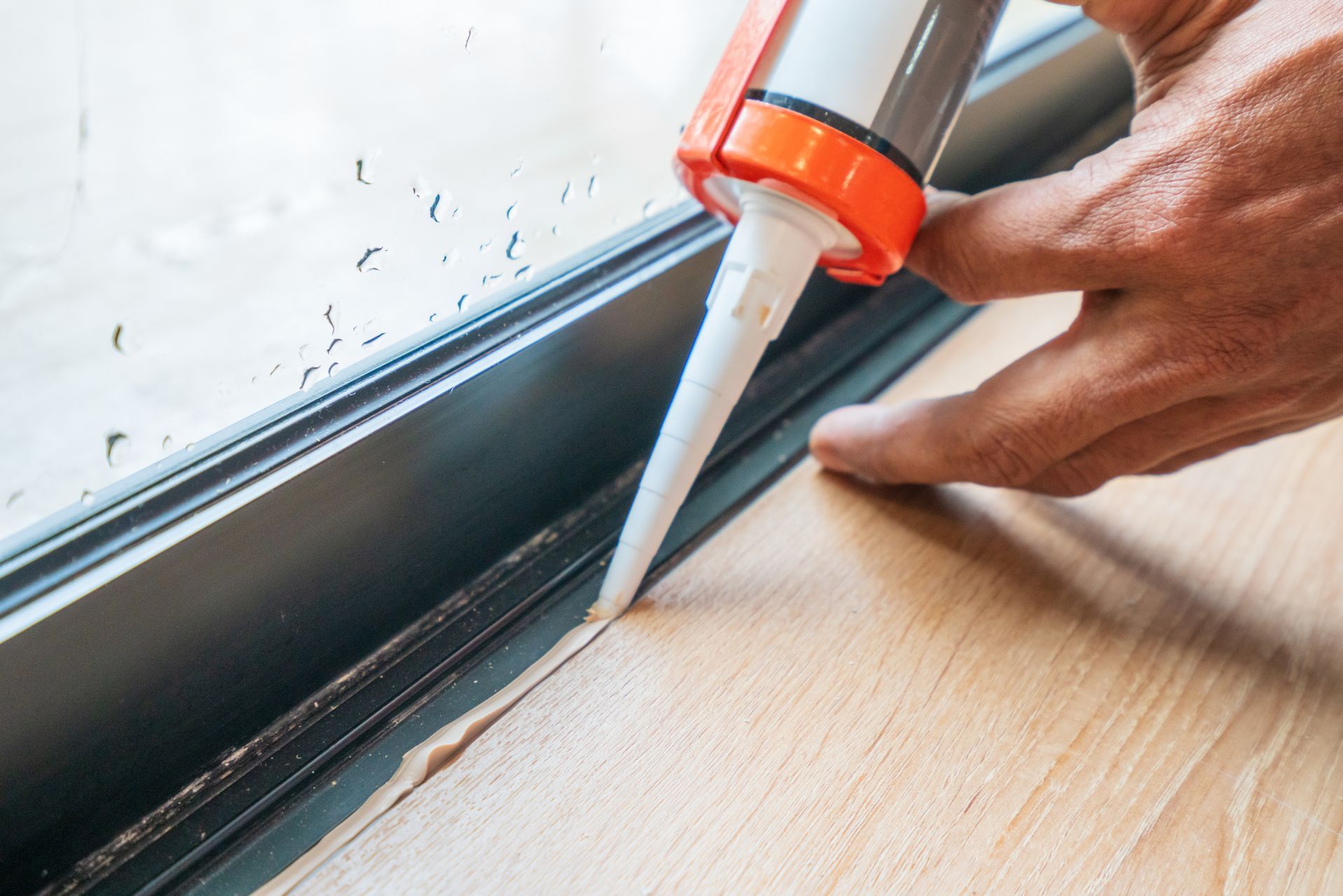 Hand applying white sealant along a window frame on a wooden floor for sealing gaps. Hand applying white sealant along a window frame on a wooden floor for sealing gaps.