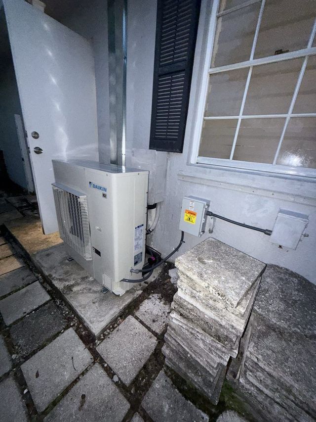 An outdoor air conditioning unit next to a window and door, with paving stones in the foreground.