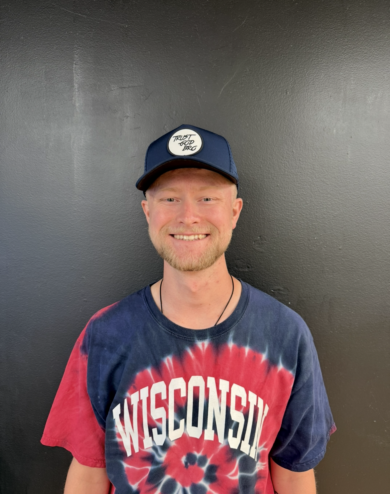 Man wearing a black hoodie and baseball cap smiles at the camera.