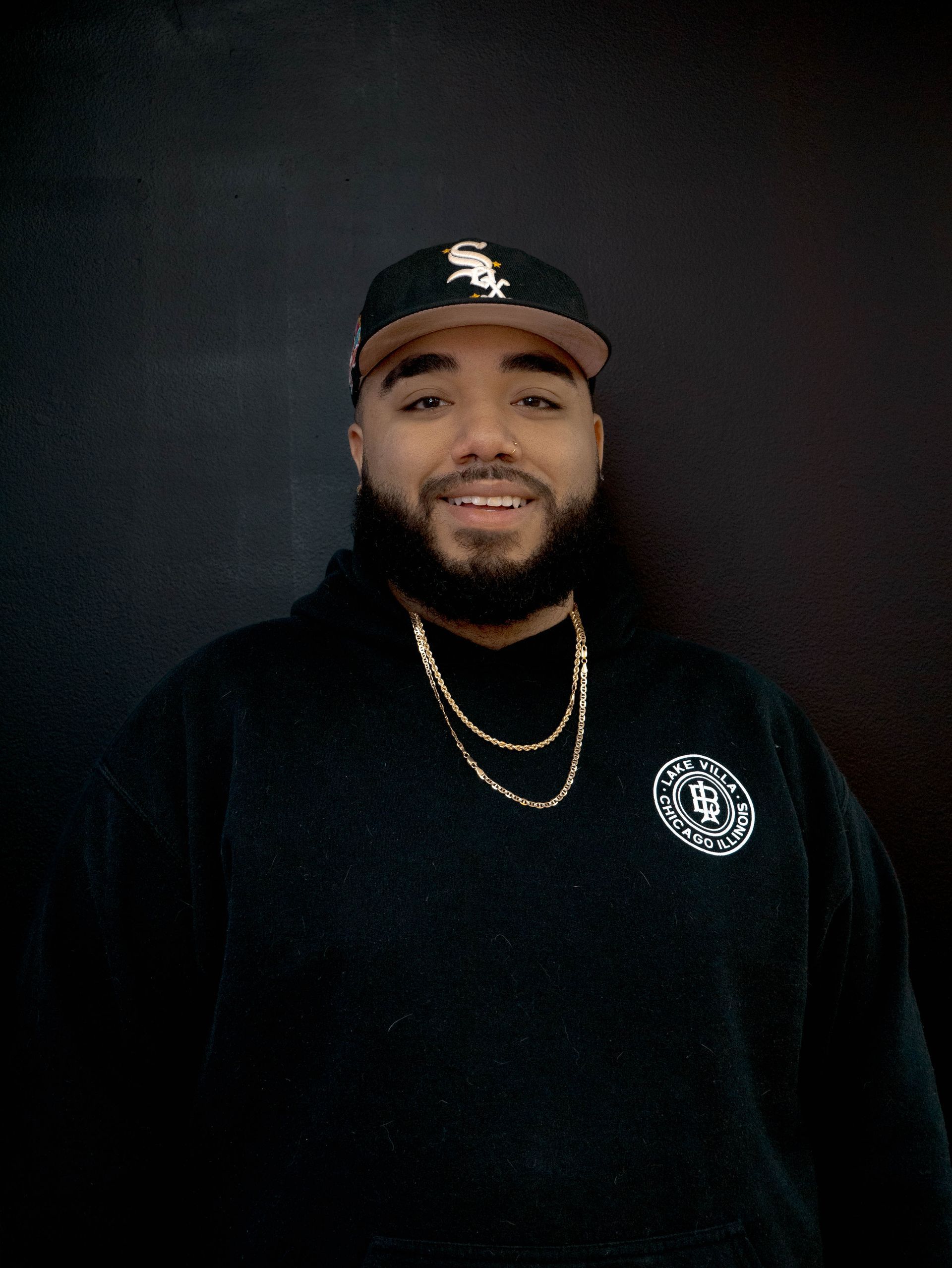 Man wearing a black hoodie and baseball cap smiles at the camera.