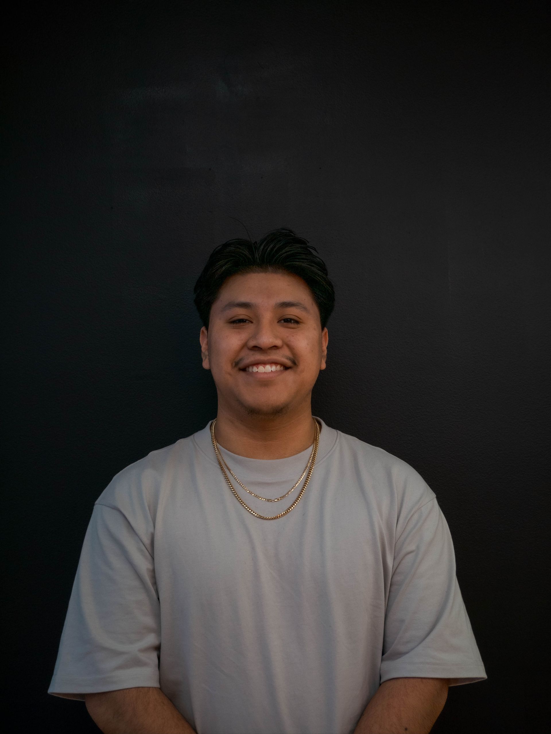 Man smiling at the camera in front of a black background, wearing a gray shirt and a gold chain.