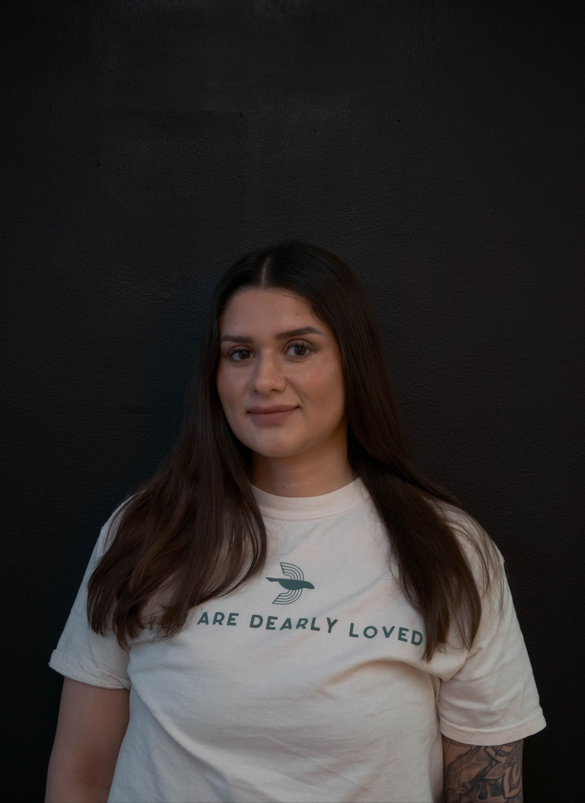 Woman in cream t-shirt with text “AGE DEARLY LOVED”, dark hair, smiling in front of a black wall.
