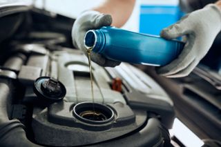 A person in gray gloves pours fresh motor oil from a blue plastic bottle into an open car engine.