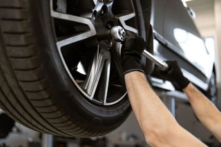 A mechanic in black gloves uses a long-handled socket wrench to tighten a lug nut on a car wheel raised in a shop.