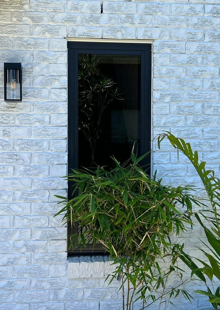 A White Brick Building With A Black Window And A Plant In Front Of It — Datco Windows & Doors Pty Ltd In Lismore, NSW