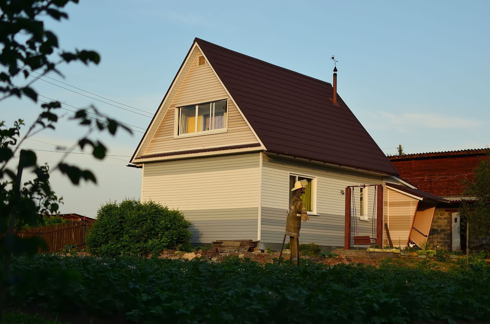 Small beige house with a red roof at sunset, partly surrounded by greenery.