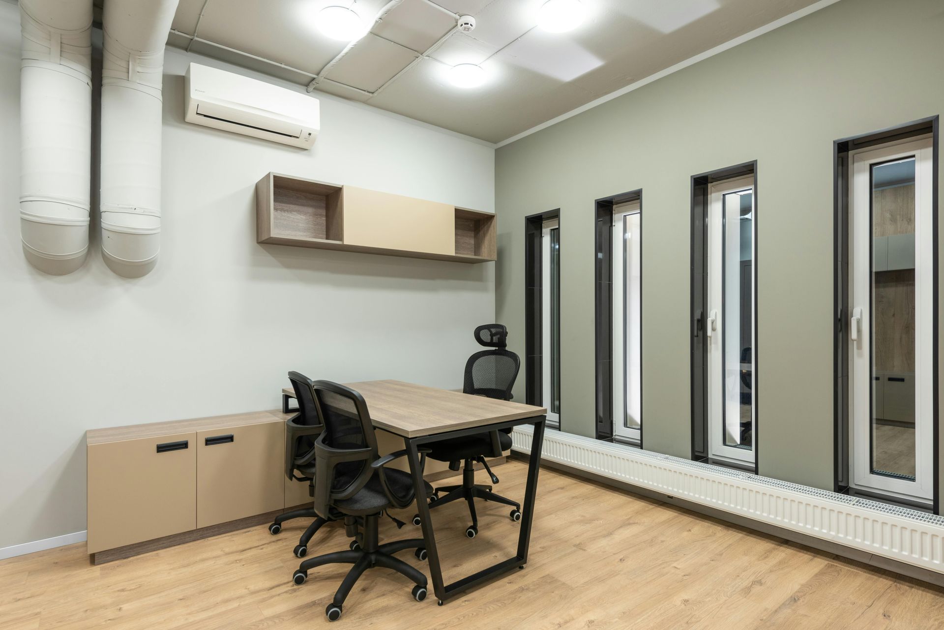 Office interior with a table, chairs, shelving, and windows. Beige and black furniture on light wood flooring.