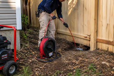Worker using a drain cleaner to unclog a pipe beside a wooden fence