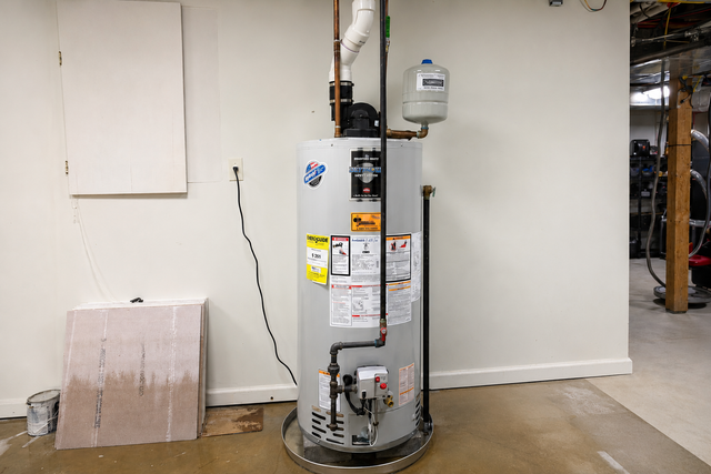 Water heater in a basement utility room, with pipes, labels, and a nearby cardboard box.