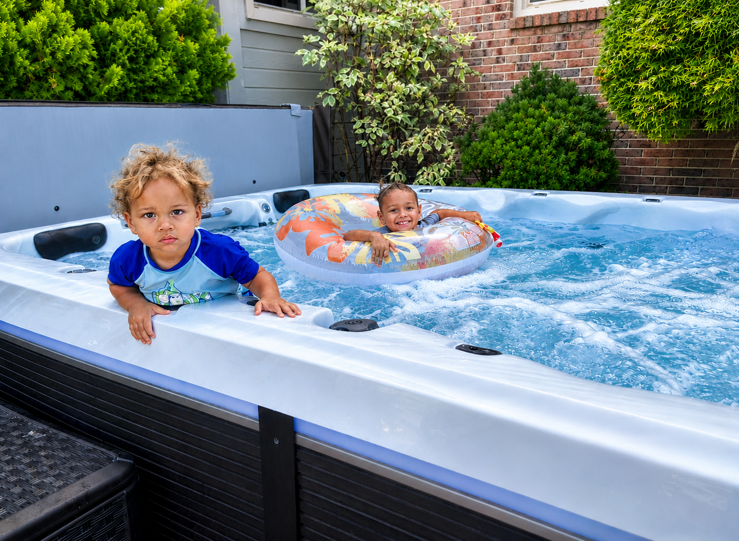 Two children playing in a backyard pool, one climbing near the edge and one floating in a ring splashing water